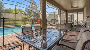 A covered outdoor area with a glass table and chairs, a pool, and a view of the surrounding landscape. - Free Baby Amenities! Stunning Disney Themed Villa! Game Room, Sunny Pool & Spa (Davenport)