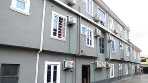 Un immeuble à plusieurs étages, doté de nombreuses unités de climatisation fixées aux murs extérieurs, d’une entrée sécurisée par un portail et d’un rez-de-chaussée peint en rouge.