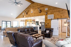Een ruime woonkamer met leren banken, een houten plafond met opgeplakte geweihanden, een keuken op de achtergrond, en een groot raam dat natuurlijk licht binnenlaat.