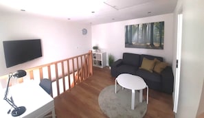 A modern living room with a flat-screen TV, a dark sofa, a small white table, and a painting of a forest.