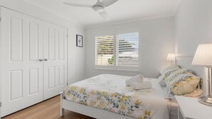 A bedroom with a bed, a bedside table with a lamp, a window with blinds, and a ceiling fan.