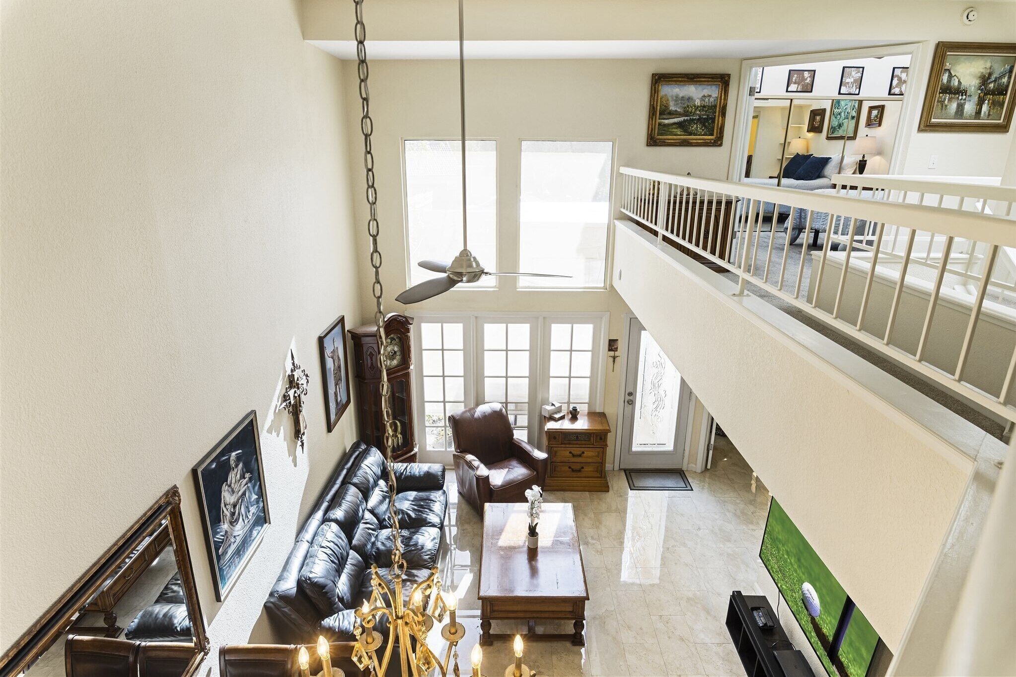 A spacious living room with a high ceiling, a ceiling fan, a black leather sofa, a wooden coffee table, and a television.