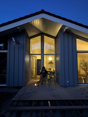 Una casa moderna con ventanas amplias, una terraza de madera y una persona sentada en una silla.