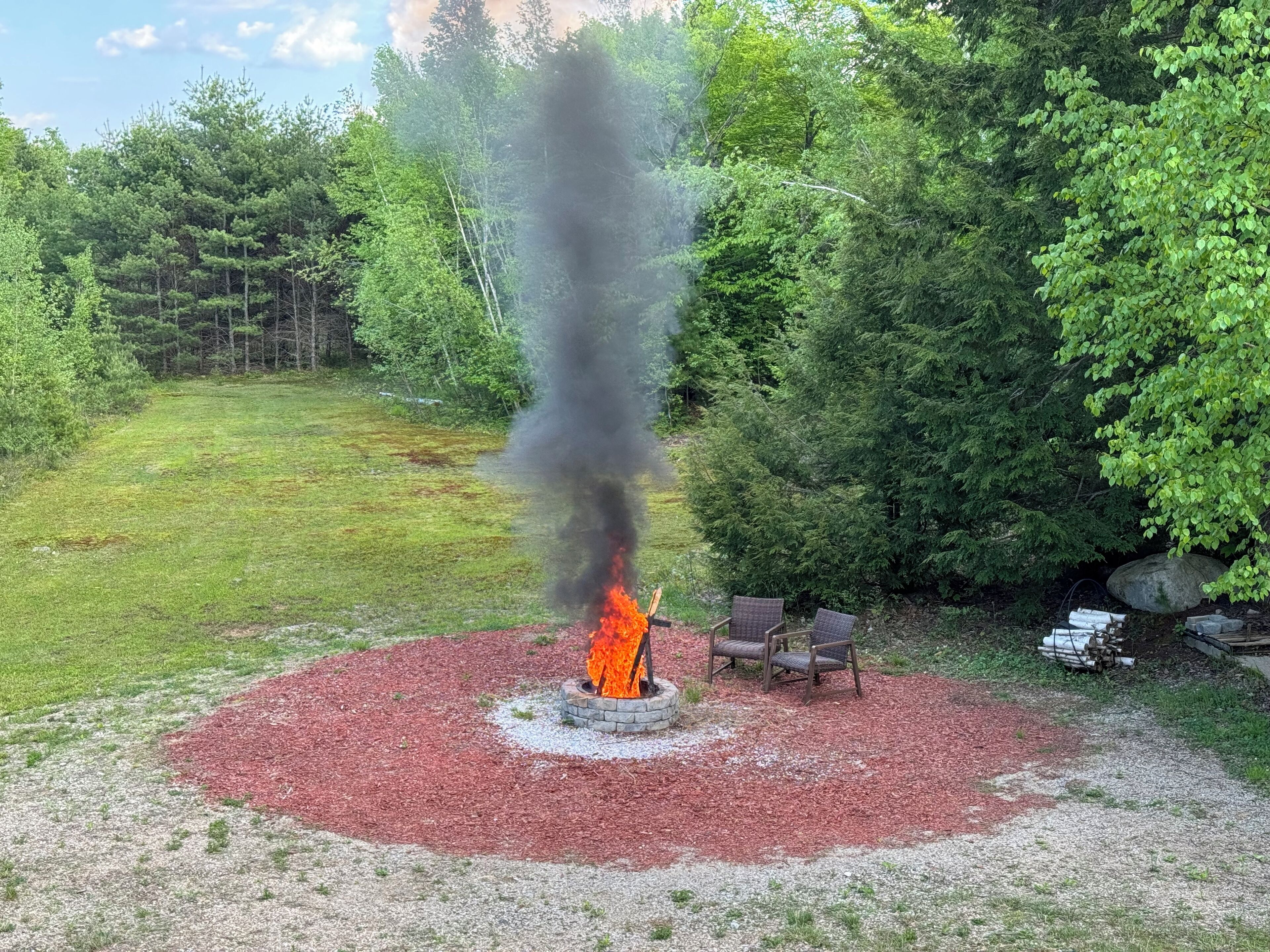 Een vuurkuil met een vlam, omringd door een rode mulchvlak, gelegen in een grasrijk buitengebied met bomen op de achtergrond.