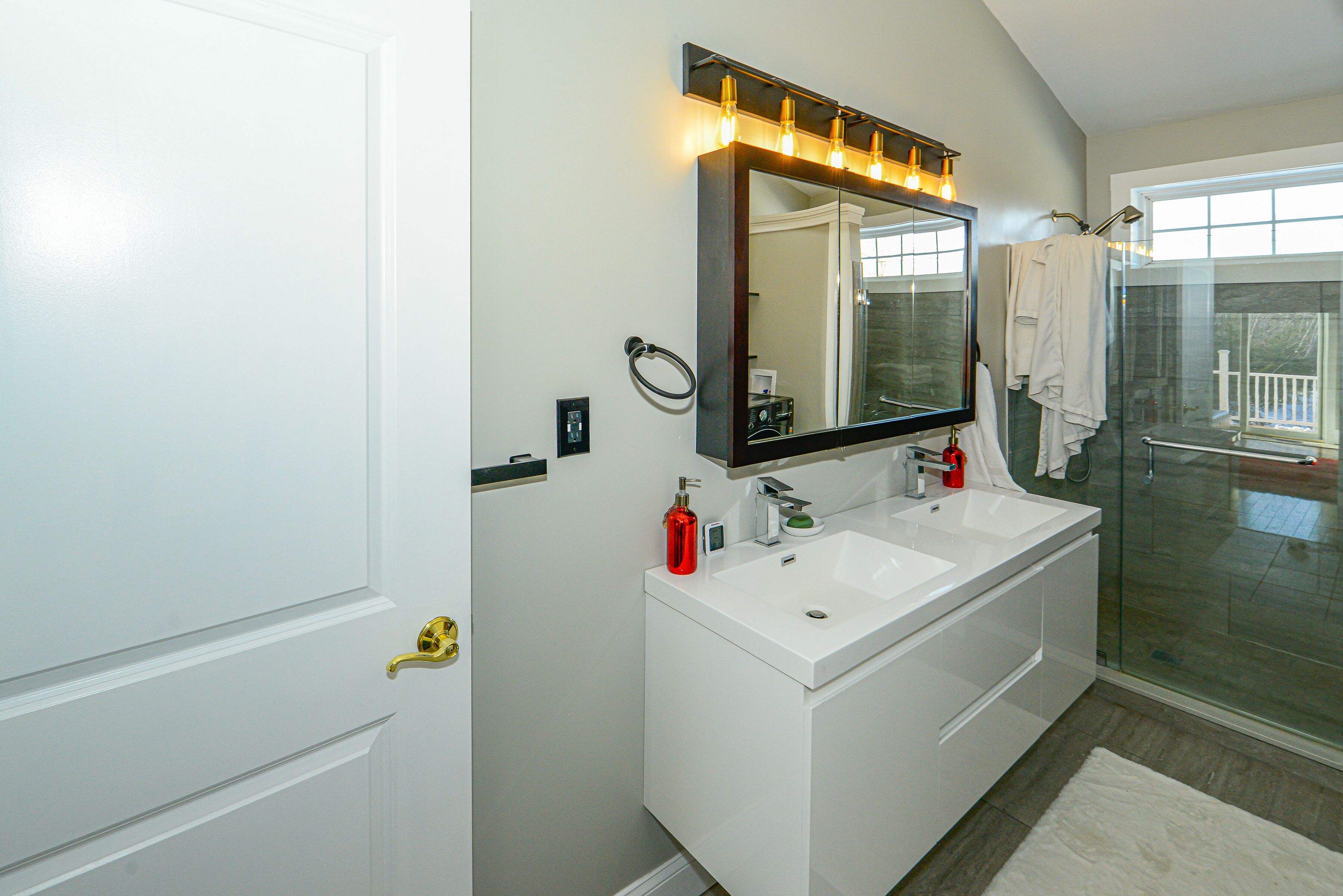 A bathroom with a double sink vanity, a large mirror with lights, a glass shower enclosure, and a white door.
