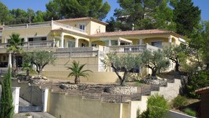 A multi-story building with balconies, a stone retaining wall, and a variety of trees including palm and olive trees. - Magnificent Gite with dominant view swimming pool and jacuzzi (L'Olivier) (Langlade)