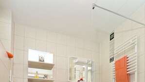 Un baño moderno con inodoro, lavamanos y zona de ducha. Hay repisas sobre el inodoro y un perchero en la pared.