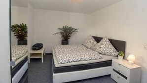 Un dormitorio ordenado con cama, mesitas de noche, lámpara y dos macetas.