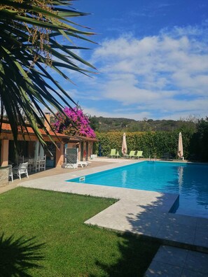 A swimming pool with lounge chairs and umbrellas, surrounded by a well-maintained lawn and a building with a red roof.