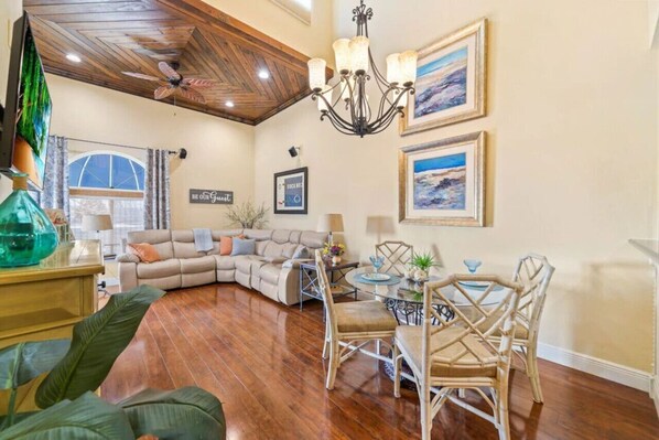 A living room with a sectional sofa, a dining table with chairs, a chandelier, and framed artwork on the walls.