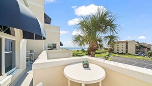Un balcón con una mesa y sillas blancas, una palmera y vistas al océano y otros edificios.