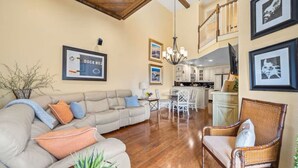 Amplia sala de estar con un sofá color crema, un sillón de mimbre y un comedor con una mesa y sillas blancas. Se ve una cocina al fondo con gabinetes blancos y una lámpara colgante.