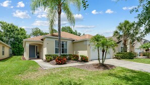 A single-story house with a front entrance, a covered porch, and a small front yard with a palm tree and flowering plants. - Perfect Getaway-4Bdrm Villa W/Pool in Gated Resort (Kissimmee)