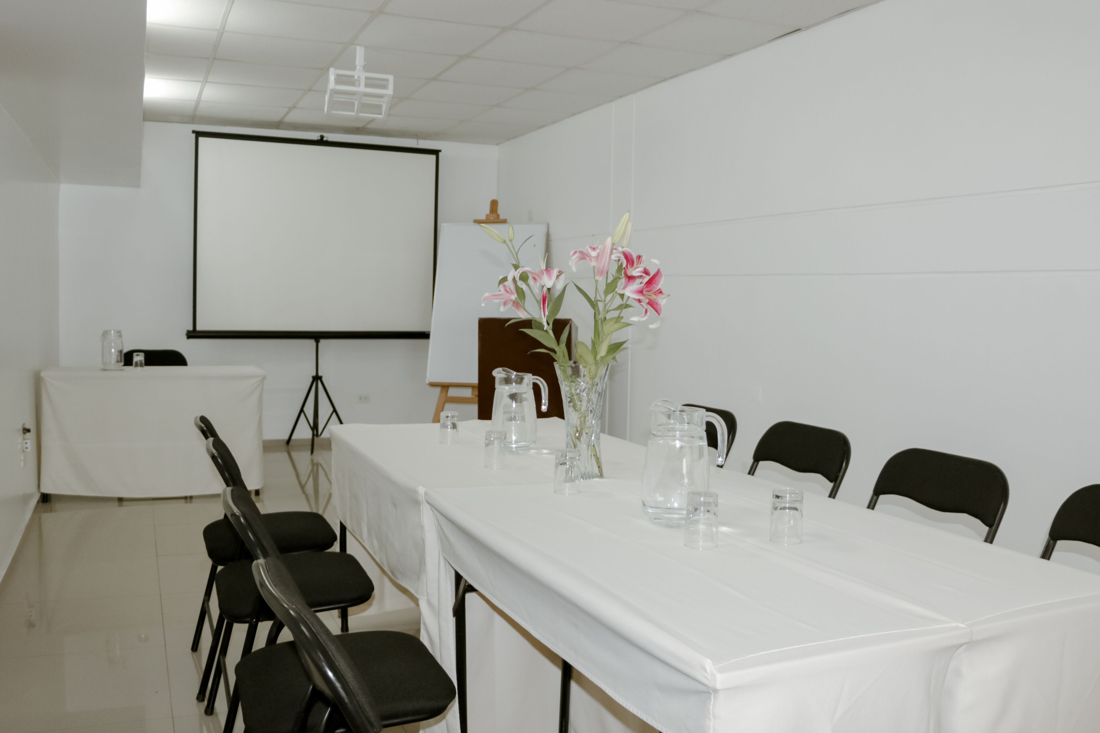 Une salle de conférence avec une longue table, des chaises, un écran de projection et un vase rempli de fleurs.