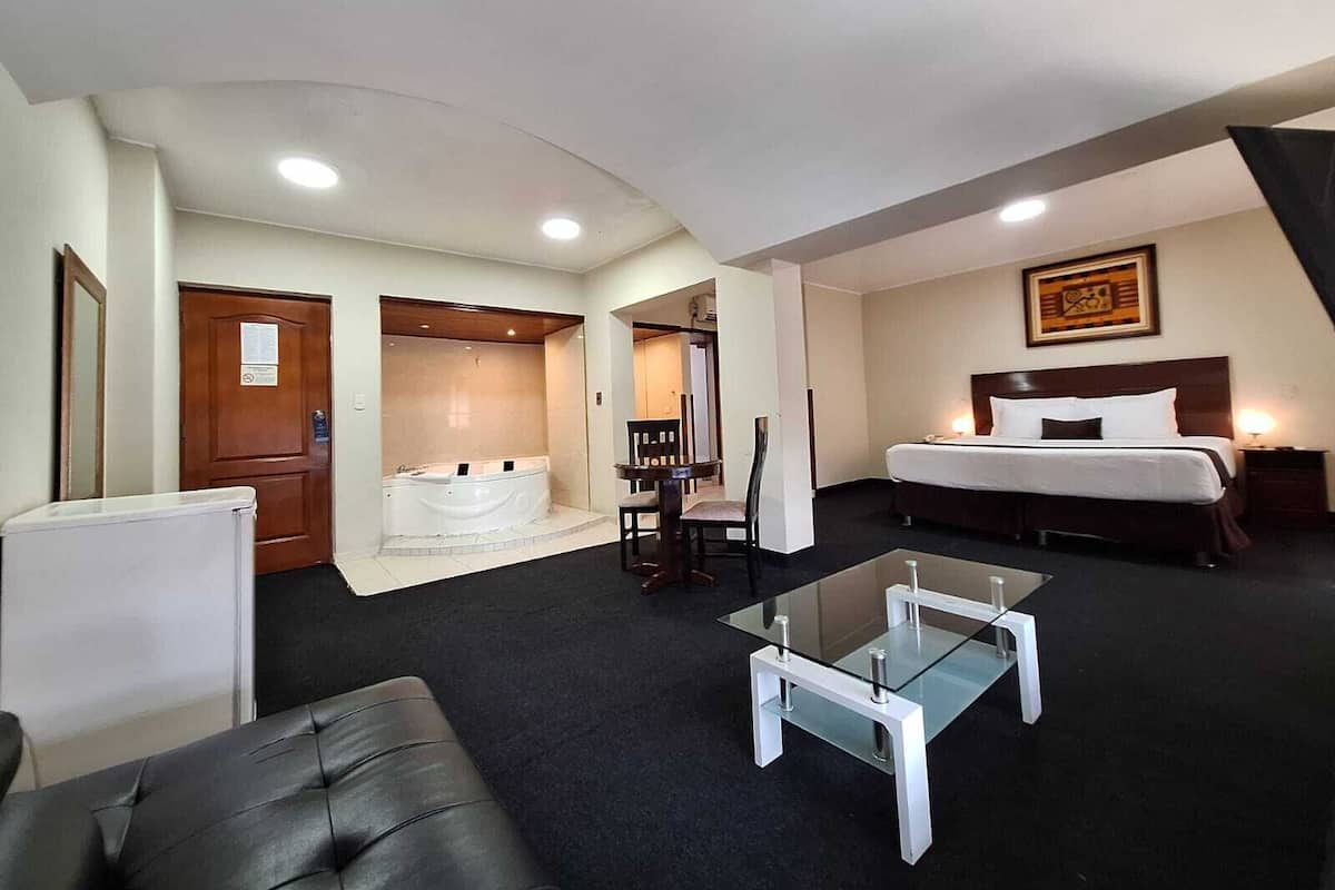 Quarto de hotel com uma cama, mesa de centro de vidro, área de estar com uma poltrona, uma mesinha, uma escrivaninha e banheiro com banheira.