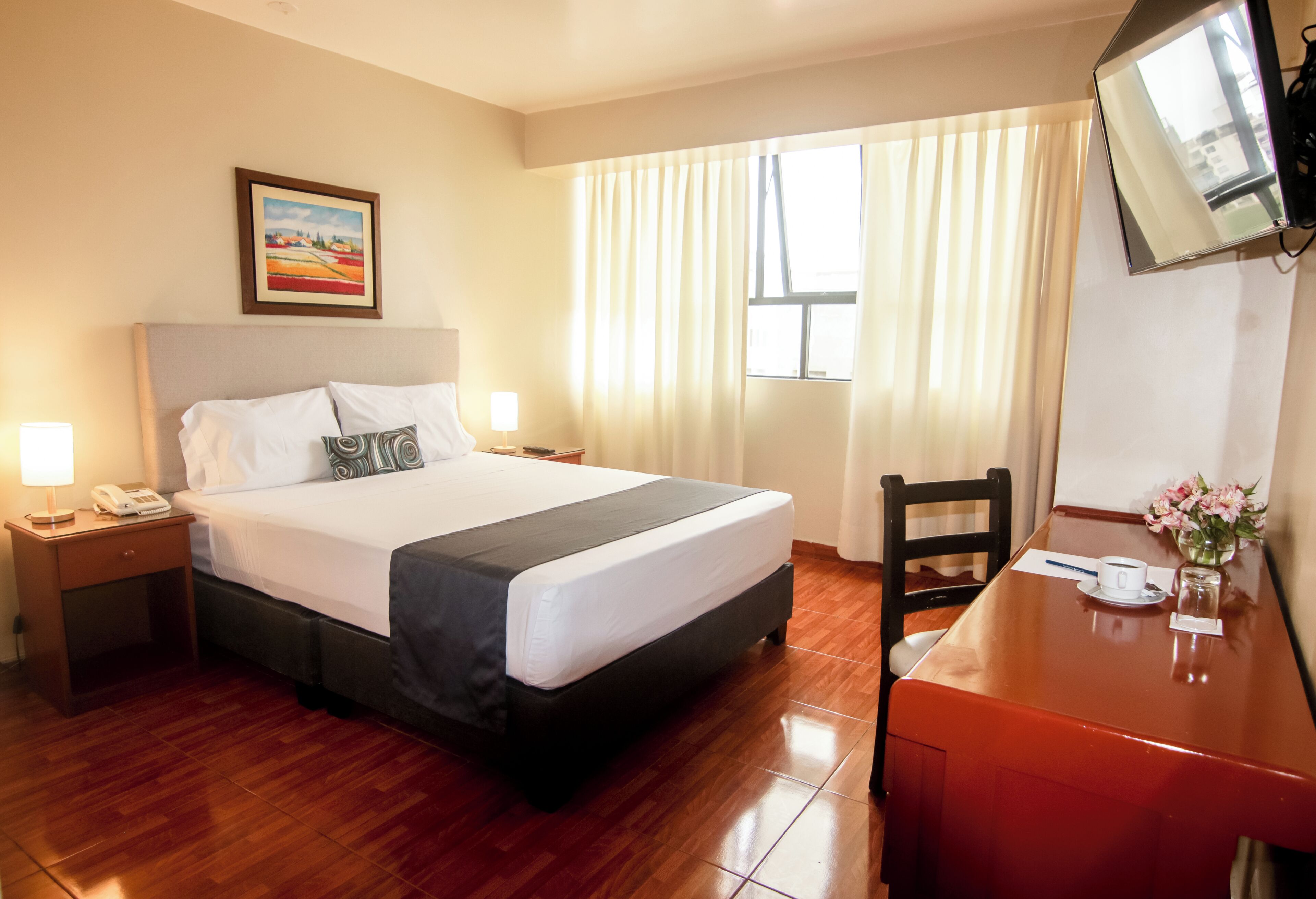 A hotel room with a bed, bedside table with a lamp, a desk with a chair, a TV mounted on the wall, and a window with curtains.