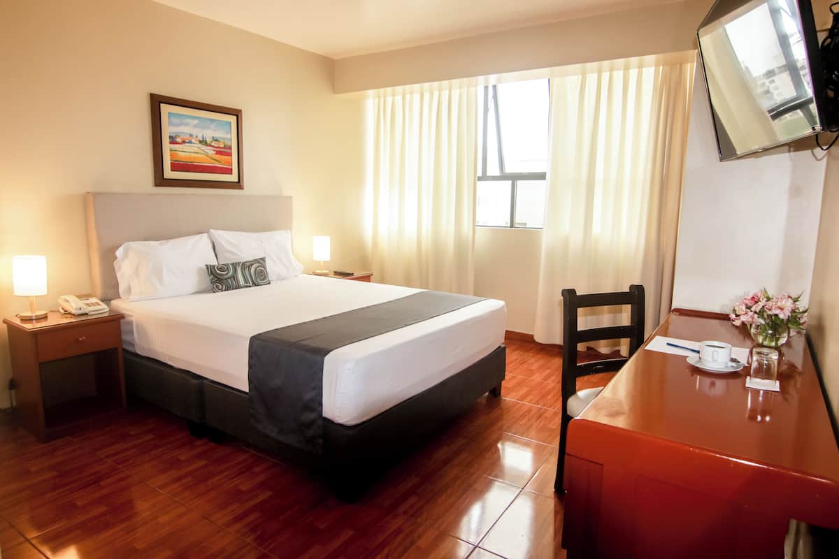 A hotel room with a bed, bedside table with a lamp, a desk with a chair, a TV mounted on the wall, and a window with curtains.