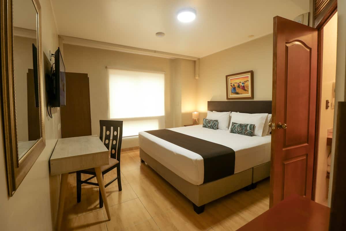 Quarto de hotel com cama, escrivaninha, cadeira, janela e porta.
