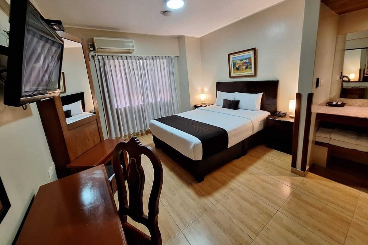 Quarto de hotel com uma cama, escrivaninha com cadeira, televisão, janela com cortinas e um quadro na parede.