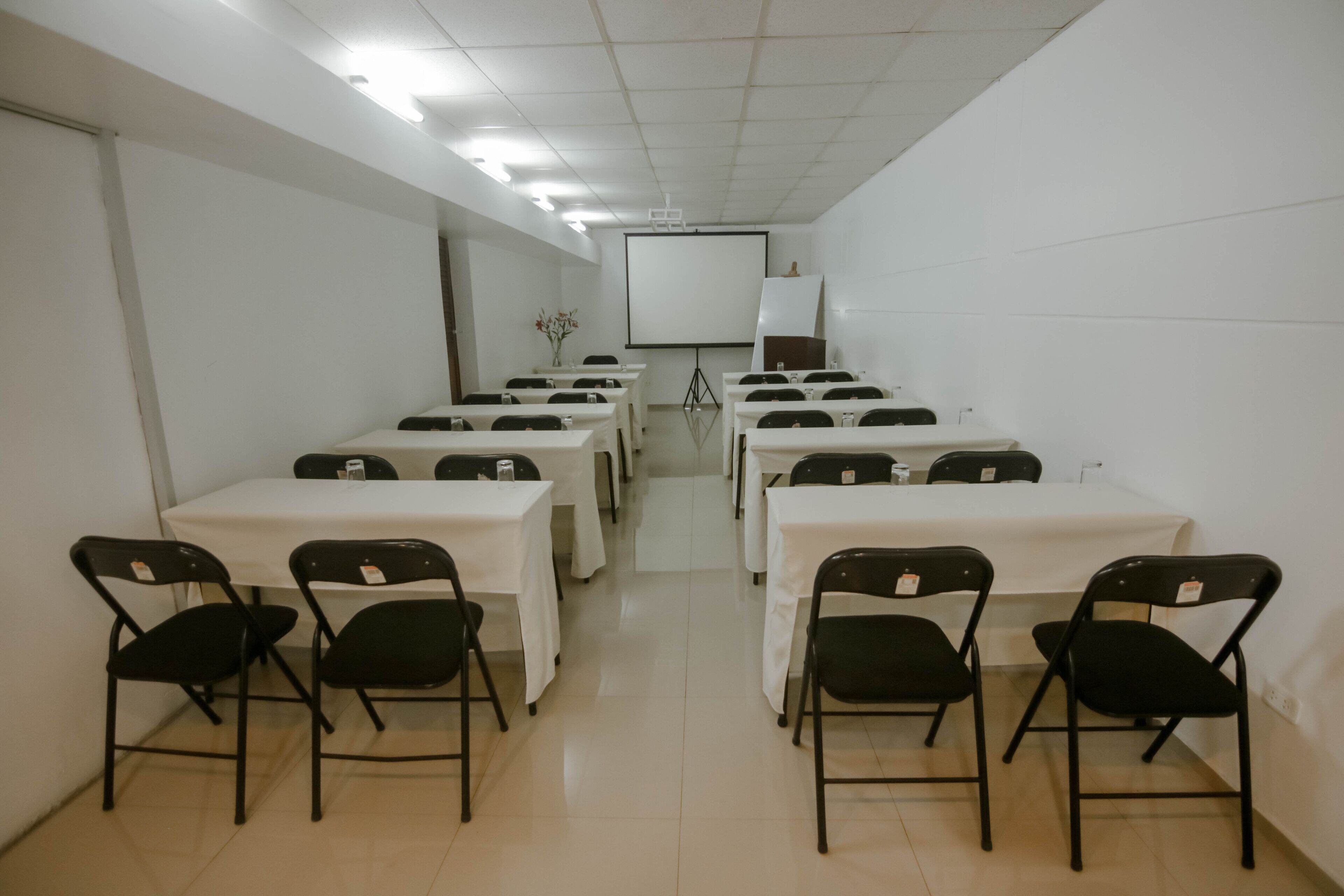 Un salón de conferencias con varias mesas y sillas, una pizarra y una pantalla de proyector.