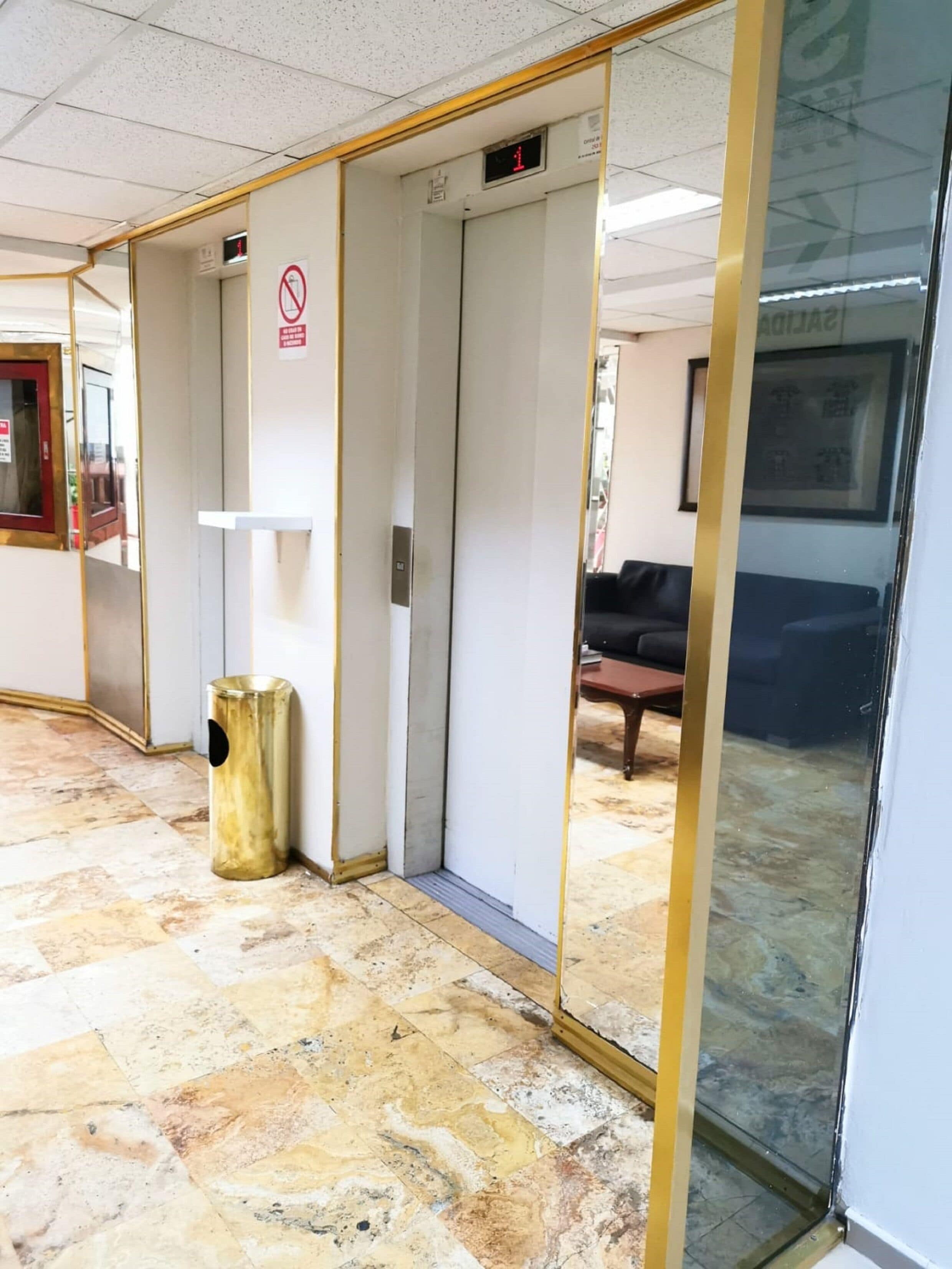 Un passadís amb sòl de rajola, un cistell de brossa, un ascensor tancat i una porta de vidre que reflecteix una habitació amb un sofà.