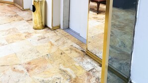 A hallway with a tiled floor, a trash bin, a closed elevator, and a glass door reflecting a room with a couch. - EPIQUS HOTEL (Lima)