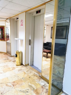 A hallway with a tiled floor, a trash bin, a closed elevator, and a glass door reflecting a room with a couch.