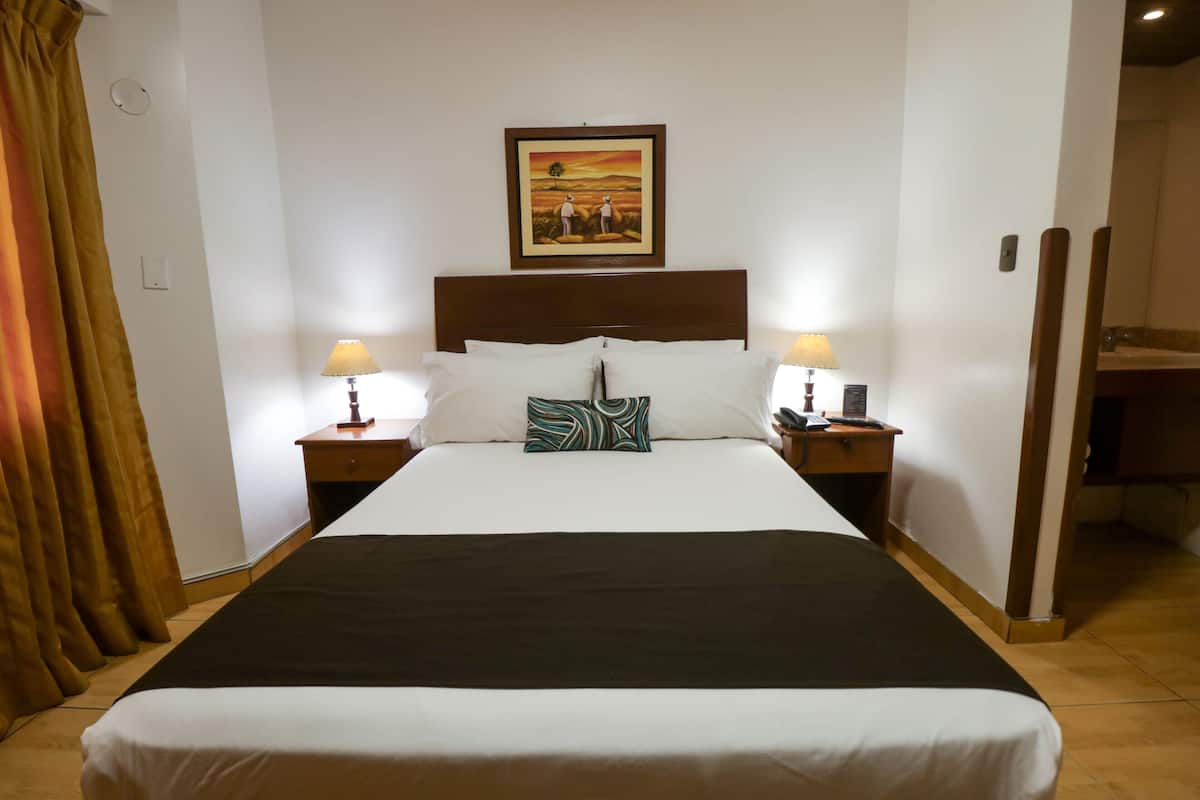 Quarto de hotel com uma cama grande, duas luminárias de cabeceira, um quadro na parede e uma cabeceira de madeira.