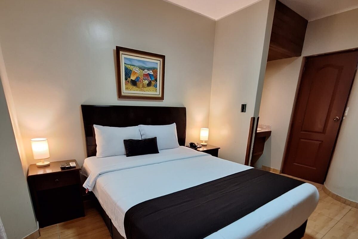 A hotel room with a large bed, two bedside tables with lamps, a wooden floor, a door, and a painting on the wall.