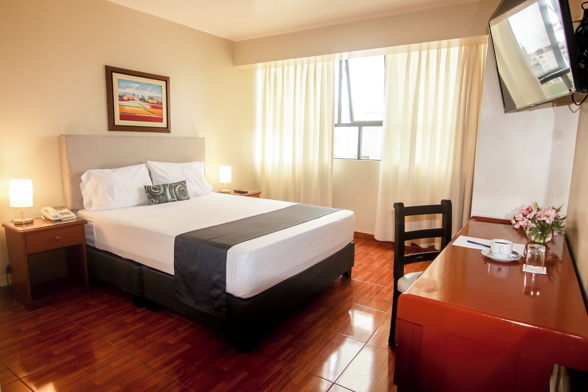 A hotel room with a bed, bedside table with a lamp, a desk with a chair, a TV mounted on the wall, and a window with curtains.
