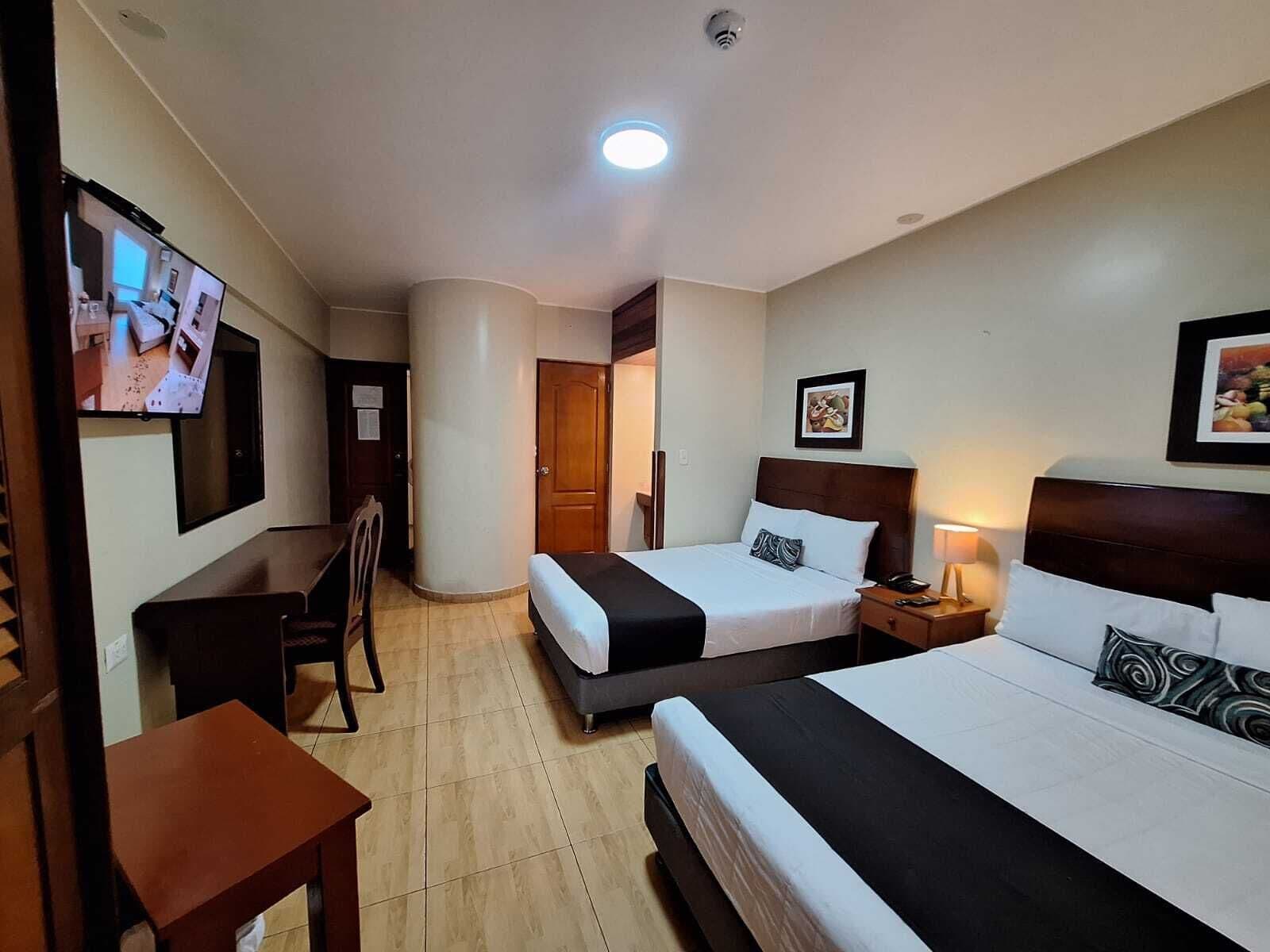 A hotel room with two beds, a desk, a chair, a TV, and a door leading to another room.