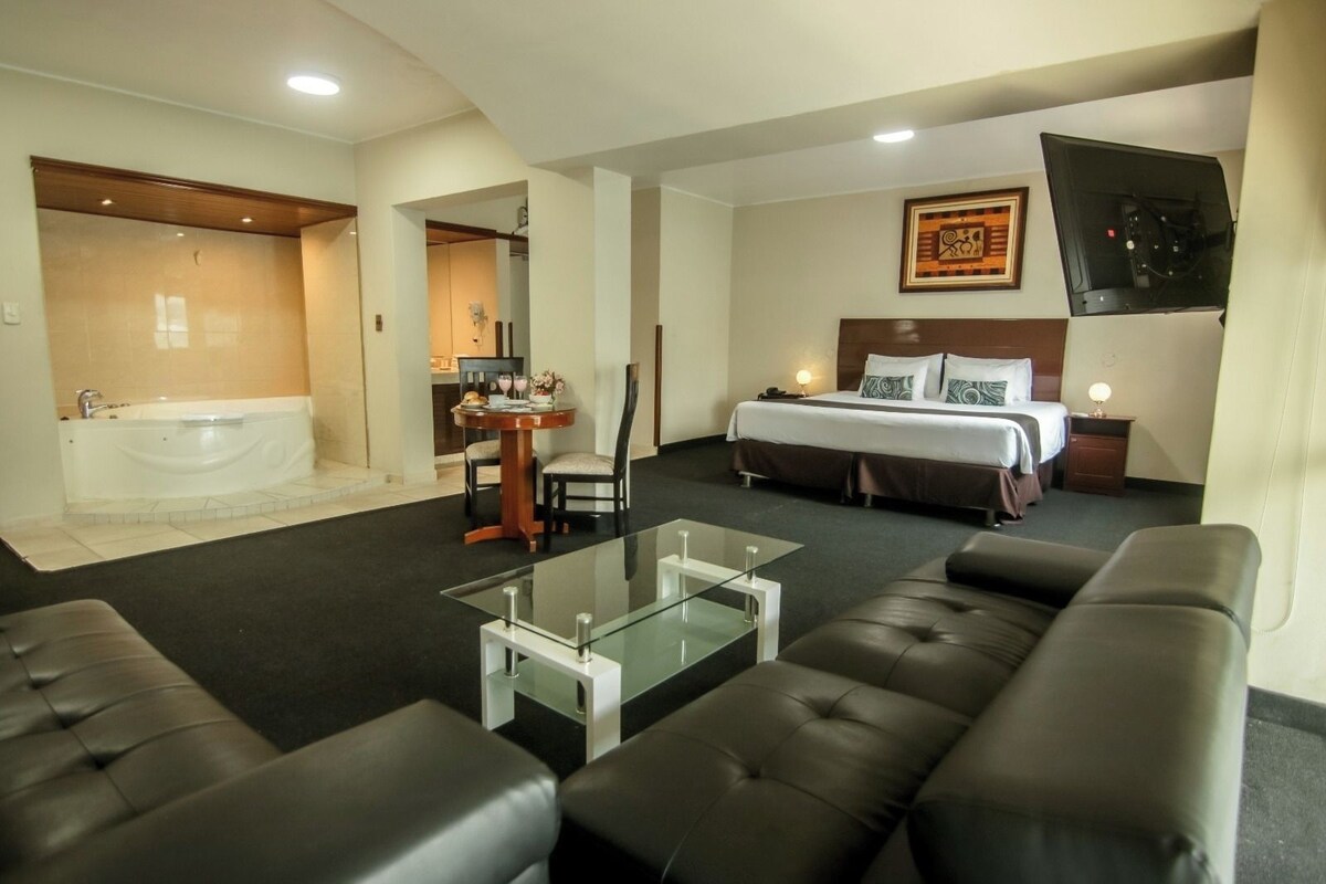 A hotel room with a bed, a sofa, a glass coffee table, a bathroom with a bathtub, and a TV mounted on the wall.