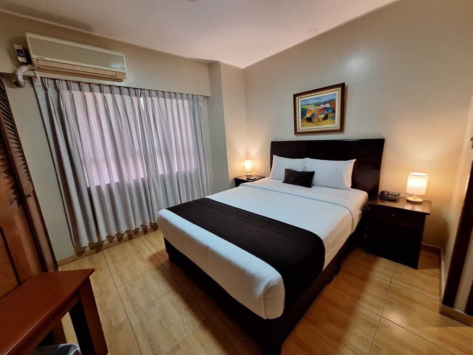 A hotel room with a bed, bedside tables, a nightstand, an air conditioning unit, curtains, and a painting on the wall.