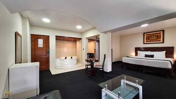 Habitación de hotel con cama, una mesa de centro de vidrio, un área de descanso con una silla, una mesita y una bañera.