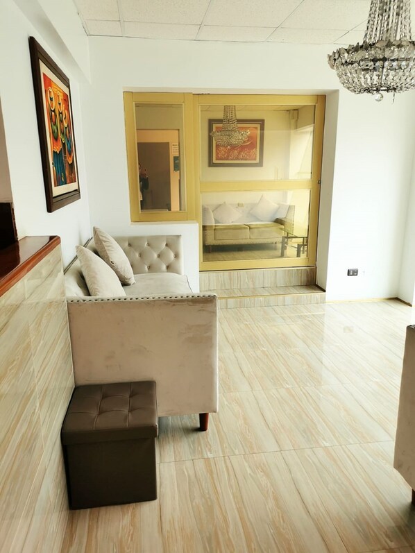 A modern living room with a beige sofa, a small ottoman, a chandelier, and a large window with a view of a balcony.