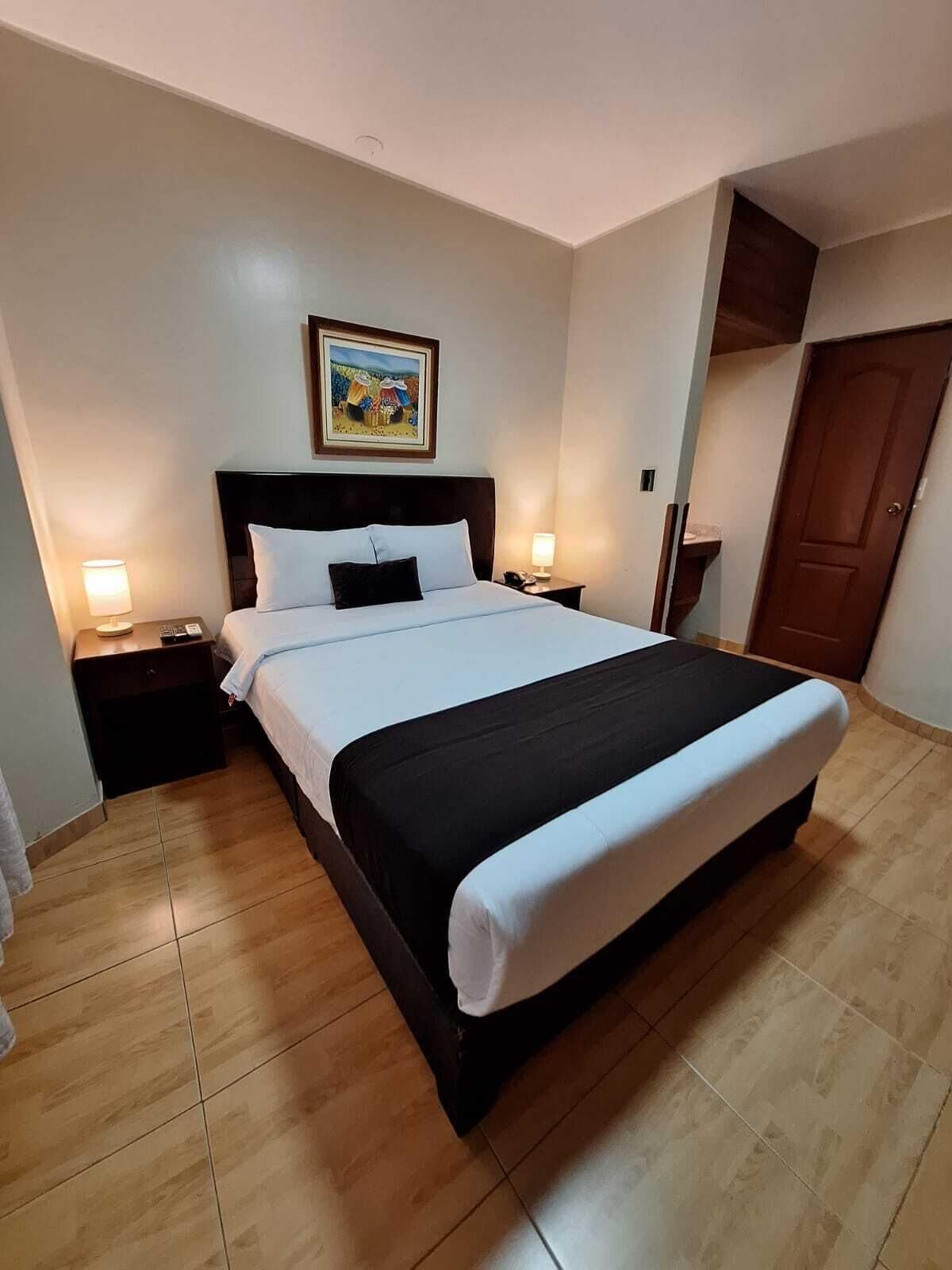 A hotel room with a large bed, two bedside tables with lamps, a wooden floor, a door, and a painting on the wall.