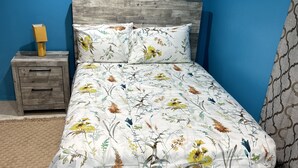 A bedroom with a wooden bed frame, floral bedding, a nightstand with a lamp, and a blue wall.