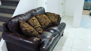 A dark brown leather sofa with patterned cushions in a room with white tiled flooring and a staircase in the background.