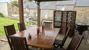 Een houten buitentafel met stoelen, een barbecue en een glazen karaf met rode drank.