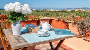 Une table en bois sur laquelle se trouvent une théière blanche, une tasse, une soucoupe, un sachet de thé, un livre et un vase avec des fleurs blanches, le tout placé sur une terrasse offrant une vue sur la mer et les bâtiments.