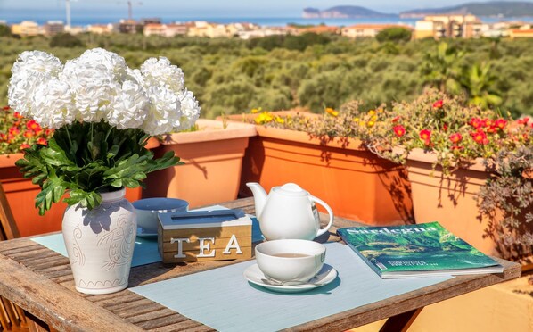 A table with a white teapot, cups, a book, and a vase with white flowers on a terrace with a view of the sea and buildings.