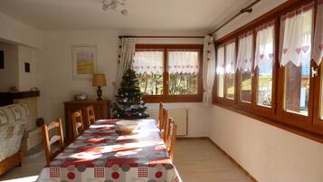 A dining room with a long table, chairs, a Christmas tree, and a window with curtains.
