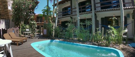 A swimming pool with wooden deck, lounge chairs, and tropical plants.