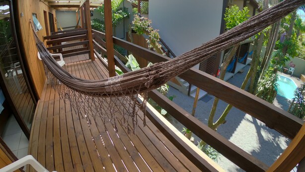 Terraza de madera con una hamaca, barandas de madera y vistas a una piscina y follaje.
