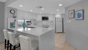 Una cocina moderna con muebles blancos, una isla central con taburetes y electrodomésticos como microondas y refrigerador.