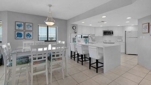 Una cocina moderna con armarios blancos, una isla central y un comedor con sillas blancas y una mesa de vidrio.