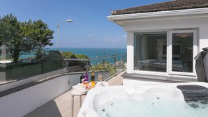 Un jacuzzi en un balcón con vista al mar, una mesa con botellas y vasos, y una sala con paredes de vidrio.