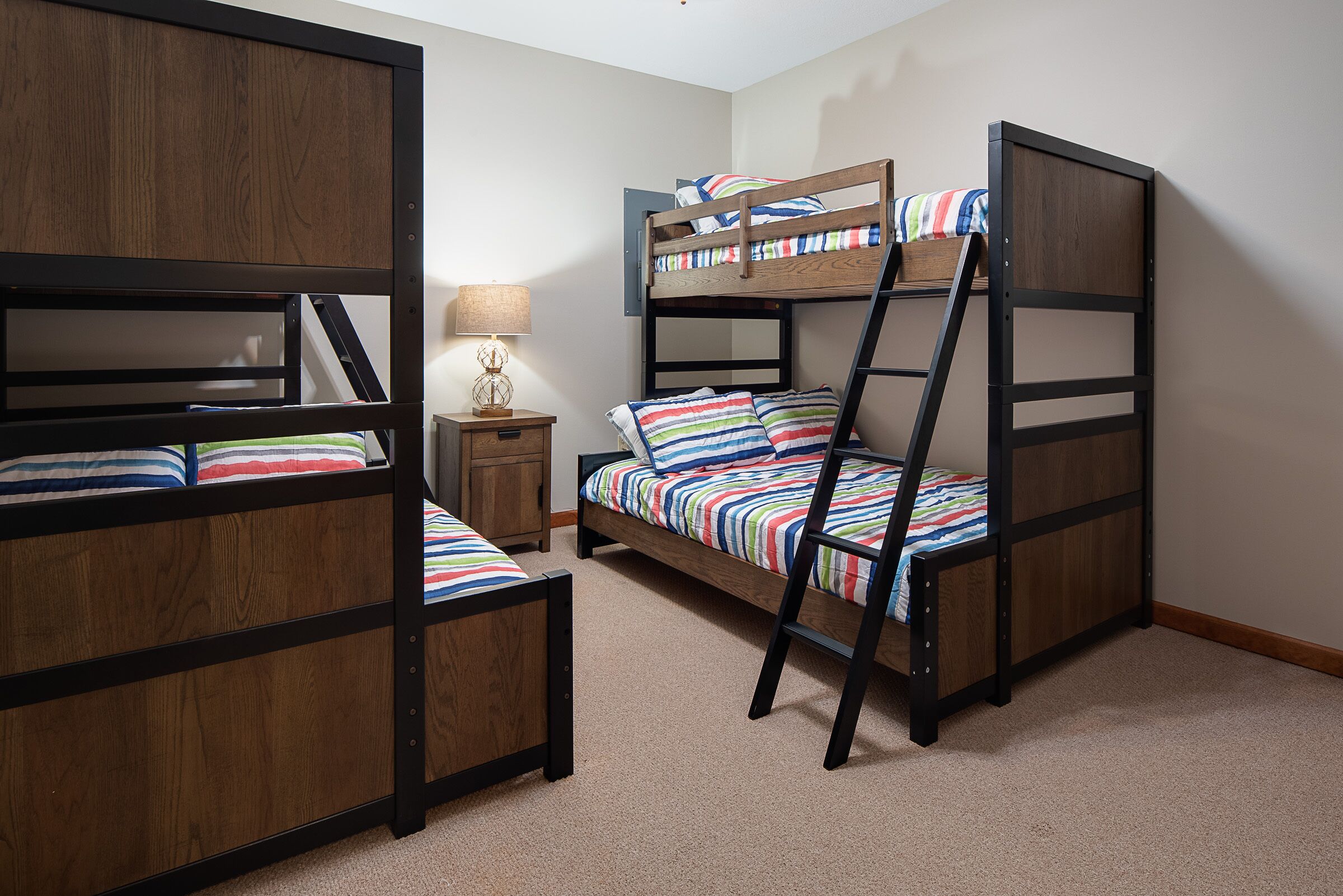 A bunk bed room with a wooden frame, a nightstand with a lamp, and a carpeted floor.