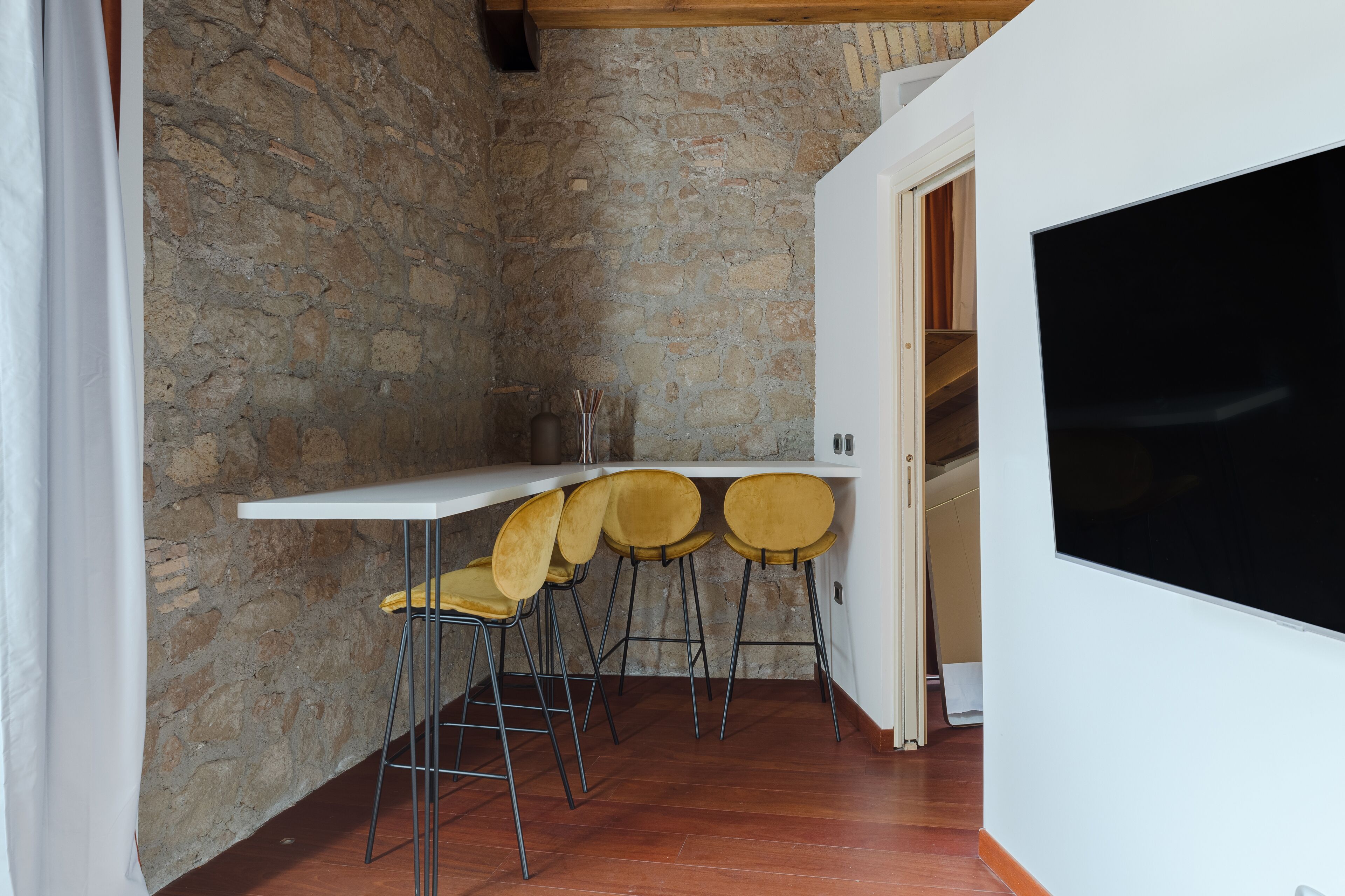 A small kitchen with a stone wall, a white countertop, and three yellow chairs.