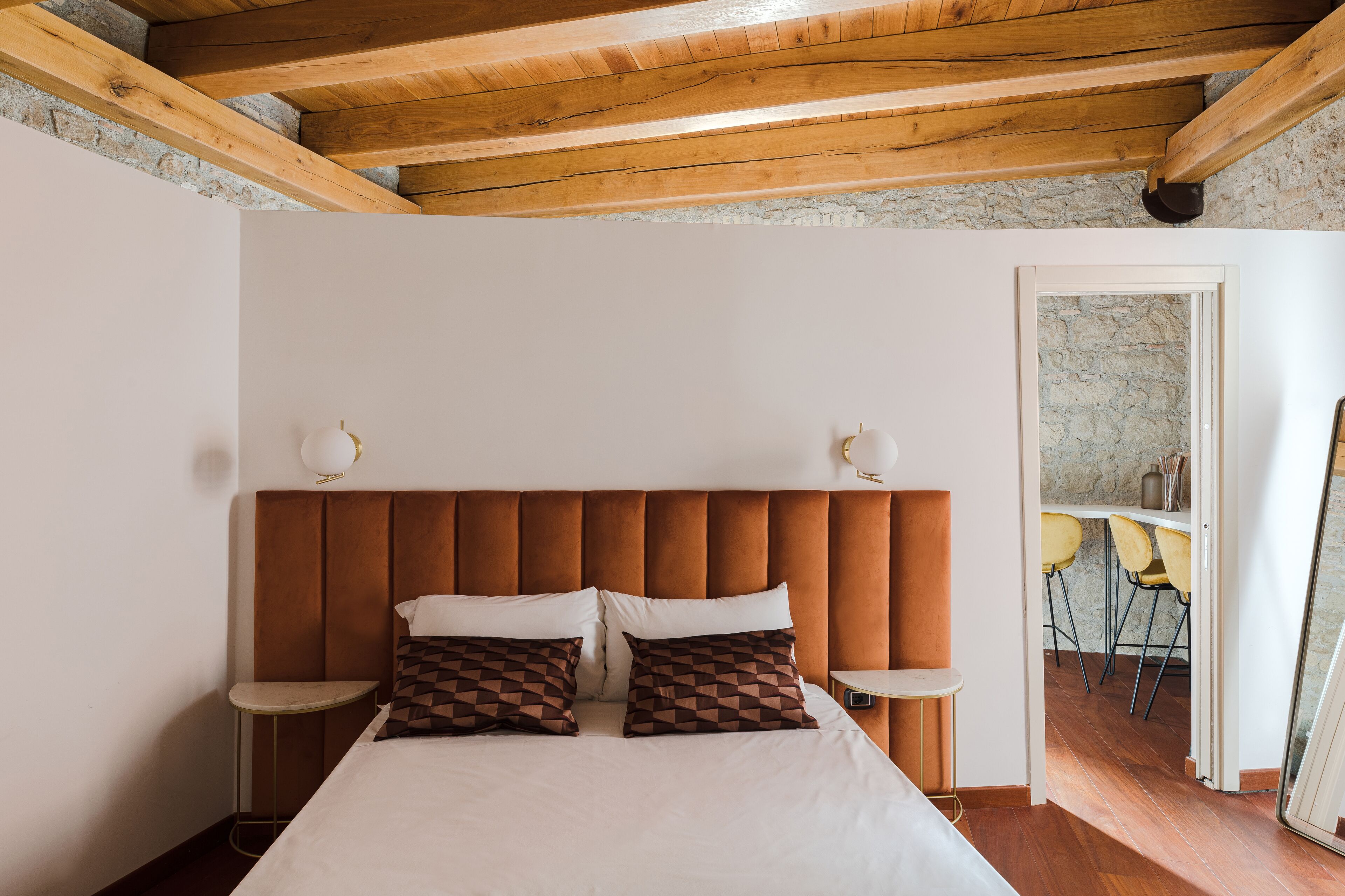 A bedroom with a wooden ceiling, a bed with white and patterned pillows, bedside tables, and a view of a dining area through an open door.